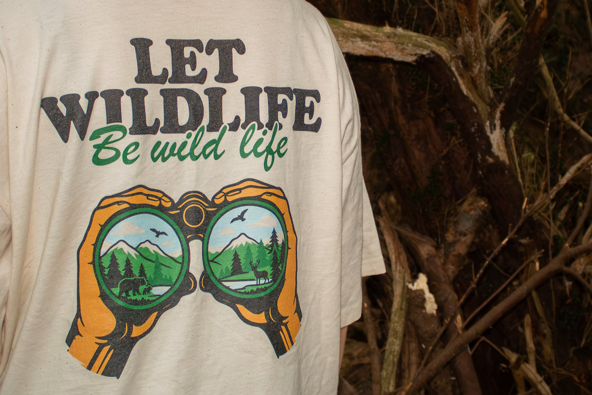 A person wearing a natural cotton cream-colored t-shirt with a graphic that reads 'LET WILDLIFE Be wild life' above a pair of binoculars showing a forest landscape. The background is a dark, wooded outdoor environment.
